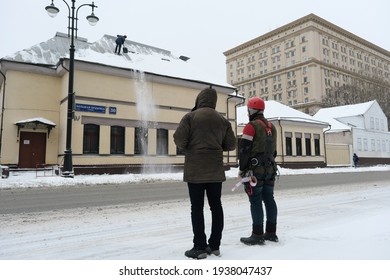 MOSCOW, FEB.25, 2021: Two Male Workers Are Watching On Snow Roof Cleaning Worker. Worker In Safety Suit With Personal Protection Equipment. Snow And Ice Removal Winter Works After Snow Blizzard, Storm