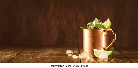 Moscow mule cocktails in copper mug with lime, ice, ginger beer, vodka and mint. Wooden background, bar tools, negative space