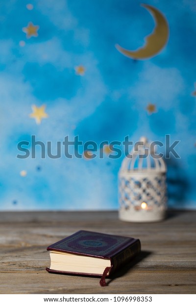 Moscow, Russia. 05/21/2018. Sacred book of\
Quran on a wooden surface. Blue night sky with crescent moon and\
candle holder in the\
background