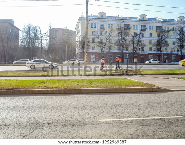 Moscow. Russia. 06 April 2021.\
Orange-uniformed janitors sweeping the lawn along a road on a city\
street. Sunny spring day. The work of the municipal public\
service.