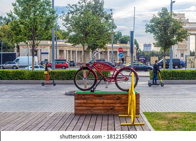 Moscow, Russia, 07/09/2020. Red Bicycle On A Wooden Pedestal, Against The Background Of The Central Entrance To Gorky Park And People Rushing Past On Scooters