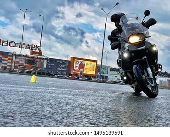 Moscow, Russia - August 18, 2019: Older Man Wearing All Black Protective Gear Is Riding A Black BMW Motorcycle Around The Cone On Parking Lot Of A Store. Very Cloudy After The Rain. Zhavoronki Globus