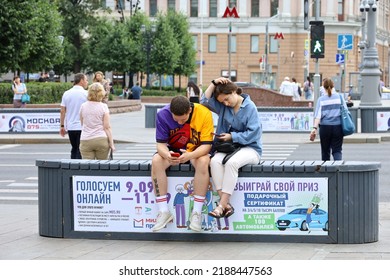 Moscow, Russia - August 2022: Couple with smartphones sitting on a street bench with advertising elections of deputies of municipalities. Online voting, leisure in summer city