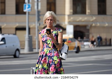 Moscow, Russia - August 2022: Girl with tattoos wearing dress with flower pattern walking with smartphone on a street. Using mobile phone in summer city