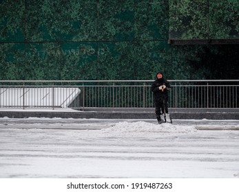 Moscow. Russia. February 12, 2021. A Utility Worker Wearing A Protective Mask Shovels Snow With A Shovel On A City Street During A Snowfall On A Winter Day.