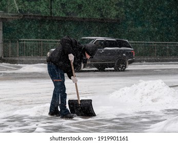 Moscow. Russia. February 12, 2021. A Utility Worker Wearing A Protective Mask Shovels Snow With A Shovel On A City Street During A Snowfall On A Winter Day.