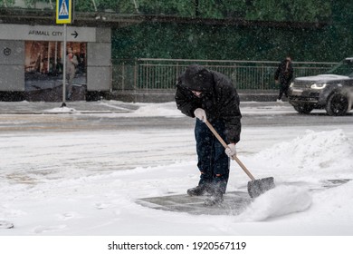 Moscow. Russia. February 12, 2021. A Utility Worker Wearing A Protective Mask Shovels Snow With A Shovel On A City Street During A Snowfall On A Winter Day.