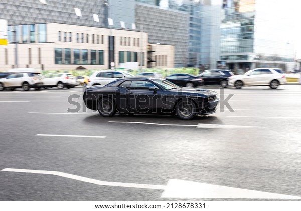 Moscow, Russia - February 2022:\
Dodge Challenger car in the motion on urban road with blurred city\
traffic background. Black muscle car is driving on the\
street