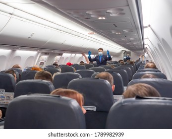 Moscow / Russia, July 2020: Steward In Mask Or Flight Attendant Shows Rules Of Behavior In Aircraft, Cabin Of The Plane, Boarding, People Sitting In Their Seats