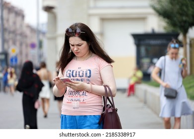 Moscow, Russia - July 2022: Concerned Woman Wearing Pink Top With Inscription Walking With Smartphone On A Street In Crowd Of People. Using Mobile Phone In Summer City