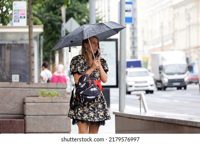 Moscow, Russia - July 2022: Girl With Umbrella Standing  On A Street And Talking On Mobile Phone On Cars Background. Rainy Weather In City, Summer Storm