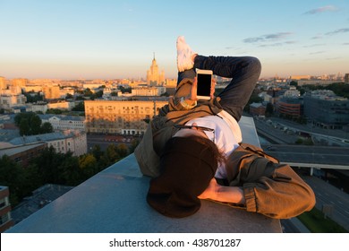 MOSCOW, RUSSIA - JUNE 13, 2016: Young and brave male sitting on the edge of high roof looking at phone similar to iphone and stunning view of city in the summer during dawn