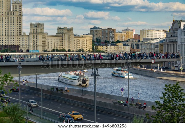 Moscow, Russia, June 16, 2019. Park Zaryadye.\
Floating bridge with people. Pleasure boats sail onMoscow River.\
Cars on track under floating bridge. Stalin\'s skyscraper on\
Kotelnicheskaya\
embankment.