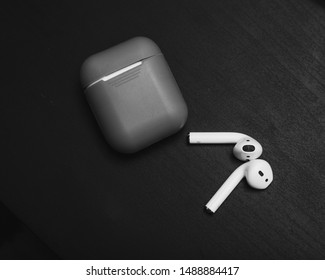 Moscow, Russia - June 2019: Wireless Bluetooth Earphones AirPods. White Earphones And Case With Grey Rubber Cover On Black Wooden Background. Selective Focus