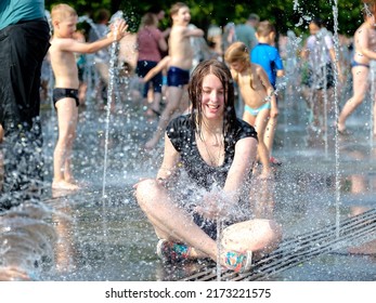 Moscow. Russia. June 26, 2021. A young beautiful smiling girl in wet clothes sits at the bottom of a city fountain and plays with water splashes. Intense heat in the city in summer.