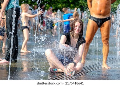 Moscow. Russia. June 26, 2021. A young beautiful smiling girl in wet clothes sits in the water jets of the city fountain and plays with splashes against the backdrop of a sunbathing man.