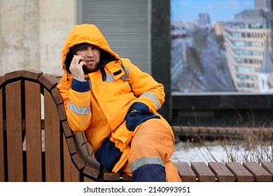 Moscow, Russia - March 2022: Public Utilities Worker Sitting On A Bench And Talking On Mobile Phone. Janitor During Rest, Street Cleaning