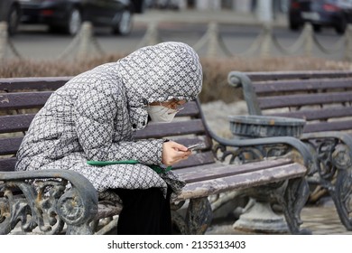 Moscow, Russia - March 2022: Woman In Face Respirator Mask Sitting With A Smartphone On The Bench In Spring City. Safety During Coronavirus Pandemic