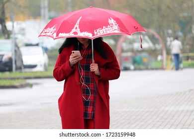 Moscow, Russia - May 2020: Rain In A Spring City, Woman With Red Umbrella Standing On A Street And Using Smartphone. Rainy Weather, Red Fashion Style