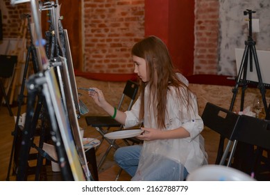 Moscow, Russia - May 26, 2022: Brown-haired Teen Girl With Paint On Hand And Cheek Drawing A Picture And Holding Paint Brush.

Young Female Painter Or Artist Painting In Front Of Easels With Canvas.