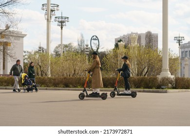 Moscow, Russia - May 3, 2022: Young Womn Went Outdoors To Ride Renting Electric Push Scooter. Moscow Public Park In Spring Day. Side View. Healthy Lifestyle. Family Walking With Baby In Baby Pram