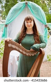 MOSCOW, RUSSIA - MAY 30, 2015: Young Woman Harp-player Participant Of Musical Band Polca An Ri Stands Holding Arpa Against Arch Decorated Fabrics. Polca An Ri Plays Traditional Irish Music.