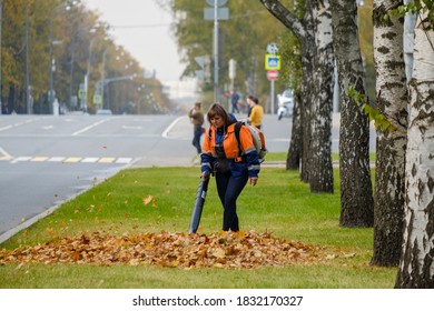 Moscow. Russia. October 11, 2020 A Woman Utility Worker Uses A Blower To Remove Fallen Leaves On A City Street. Technological Equipment. Yellow Leaves Are Flying In The Air. Seasonal Work Concept.