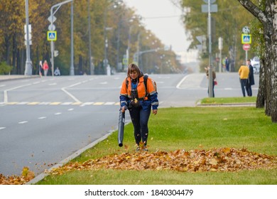 Moscow. Russia. October 11, 2020 A Woman Utility Worker Uses A Blower To Remove Fallen Leaves On A City Street. Technological Equipment. Yellow Leaves Are Flying In The Air. Seasonal Work Concept.