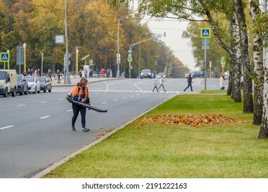 Moscow. Russia. October 11, 2020 A Woman Utility Worker Uses A Blower To Remove Fallen Leaves On A City Street. Technological Equipment. Yellow Leaves Are Flying In The Air. Seasonal Work Concept.