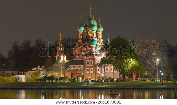 Moscow, Russia - October 13, 2021: Moscow calm\
cityscape in the autumn night. Church of the Life-Giving Trinity in\
Ostankino. Road is covered by parked cars. Electrical street\
lighting.