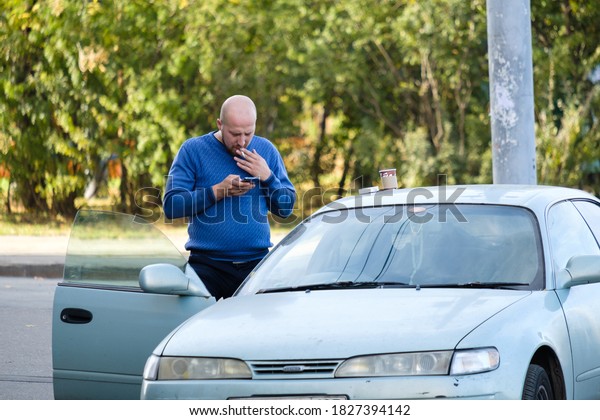 Moscow. Russia. On October 4, 2020. A bald man\
stands outside the open door of a car and smokes a cigarette while\
looking at his smartphone screen. Sunny autumn day. Tobacco smoke\
poisons the body.