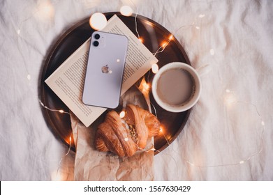 Moscow/Russia - November 22, 2019: Iphone 11 In Purple Color On Open Book With Cup Of Coffee With Fresh Croissant On Wooden Tray In Bed Over Christmas Lights. 