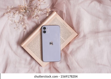 Moscow/Russia - November 22, 2019: Iphone 11 In Lilac Coloron Open Book With Flower In Bed Close Up. Selective Focus. 