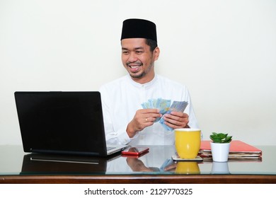 Moslem Asian man smiling while counting his money in front of laptop