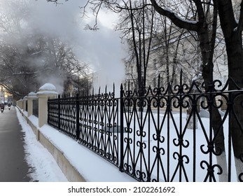 Mosow, Russia- December 23, 2021: Sidewalk And Iron Fence In Winter With Steam In The Background. White Steam From A Burst Hot Water Pipe In Winter. 