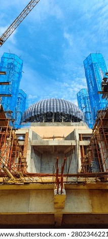 Mosque under construction, low angle view of building site with main dome, scaffolding, concrete structure, steel reinforcement and construction crane. Bright blue sky, development of Islamic religious building in Indonesia.