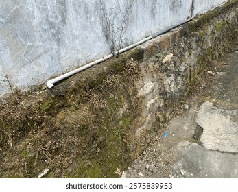 A mossy stone wall with a white pipe running along it shows signs of age and nature reclaiming the space Perfect for texture and architectural photography