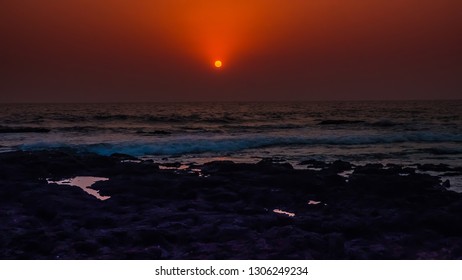 Most beautiful sunset at the sea in the northern Goa at Anjuna beach, India. The full sun of orange color sets behind the Arabian Sea with waves and rocks. Stunning tropical and exotic landscape.
