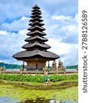 The most complete and iconic wide-angle view of Pura Ulun Danu Bratan, showing the full temple complex situated at the edge of Lake Bratan. The 11-tiered main meru tower dominates the center, flanked by a smaller two-tiered meru to the left and various ornamental shrines and statues lining the stone-paved temple courtyard. The foreground features a tranquil pond covered in green lily pads, colorful tropical flowers, a decorative frog sculpture, and stone pathway. The lake stretches behind with lush green forested hills. Dramatic cumulus clouds fill a blue sky, perfectly reflecting the moody tropical highland atmosphere.
