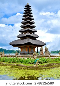 The most complete and iconic wide-angle view of Pura Ulun Danu Bratan, showing the full temple complex situated at the edge of Lake Bratan. The 11-tiered main meru tower dominates the center, flanked by a smaller two-tiered meru to the left and various ornamental shrines and statues lining the stone-paved temple courtyard. The foreground features a tranquil pond covered in green lily pads, colorful tropical flowers, a decorative frog sculpture, and stone pathway. The lake stretches behind with lush green forested hills. Dramatic cumulus clouds fill a blue sky, perfectly reflecting the moody tropical highland atmosphere.