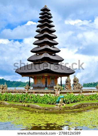 The most complete and iconic wide-angle view of Pura Ulun Danu Bratan, showing the full temple complex situated at the edge of Lake Bratan. The 11-tiered main meru tower dominates the center, flanked by a smaller two-tiered meru to the left and various ornamental shrines and statues lining the stone-paved temple courtyard. The foreground features a tranquil pond covered in green lily pads, colorful tropical flowers, a decorative frog sculpture, and stone pathway. The lake stretches behind with lush green forested hills. Dramatic cumulus clouds fill a blue sky, perfectly reflecting the moody tropical highland atmosphere.