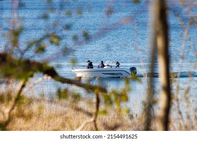 Motala Sweden May 2022
Men In A White Motor Boat Or Speedboat Fishing Seen Through Trees And Bransches And Reed In Foreground