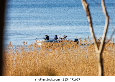 Motala Sweden May 2022
Men In A White Motor Boat Or Speedboat Fishing Seen Through Trees And Bransches And Reed In Foreground