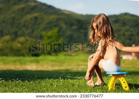 A mother applies sunscreen to her little daughter's back before the child goes for a walk. The child is sitting on a chair and green grass. Free space
