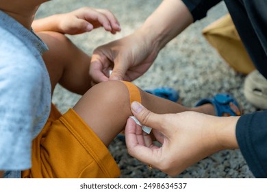 A mother is applying a band-aid to a scrape on her child's leg, who fell while playing in the park, first aid concept - Powered by Shutterstock