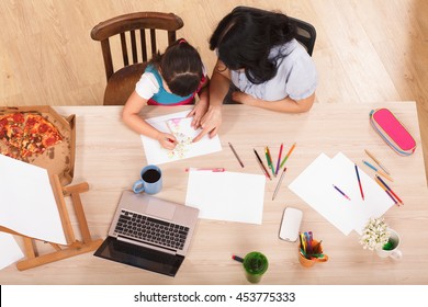 Mother Assisting Her Little Daughter In Drawing Pictures While Resting At Home. Top View Of Mother And Kid Sitting At Table In Front Of Laptop Computer.