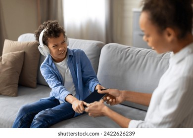 Mother attempts to take tablet from her young son who seems reluctant to give it up, while wearing headphones in modern living room setting - Powered by Shutterstock