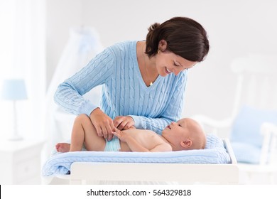 Mother and baby change diaper after bath in white nursery with bed and rocking chair. Little boy on changing table in clean dry nappy. Mom taking care of infant child. Kids room interior and hygiene