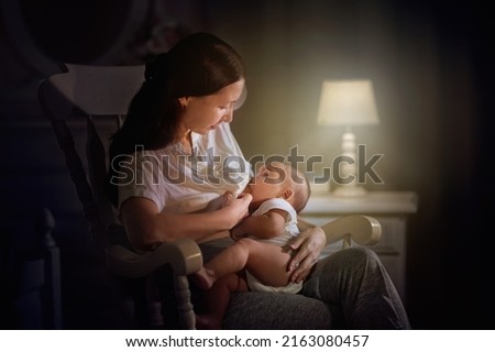 Mother and baby in dark bedroom. Asian mom with newborn boy in white rocking chair at night. Breastfeeding and infant care. Happy family at home. Bedtime and midnight feeding.