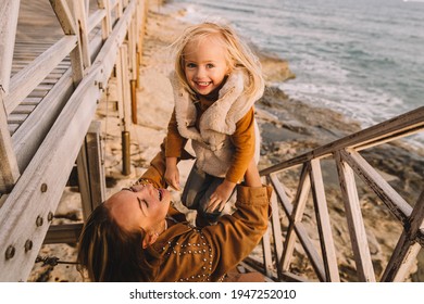 Mother With Baby Daughter Next To The Sea Smiling. Baby Girl With Blonde Hair, In A Warm Fur Vest. Spring Time, Sun Is Shining. Travel Abroad. Happy Young Family. 